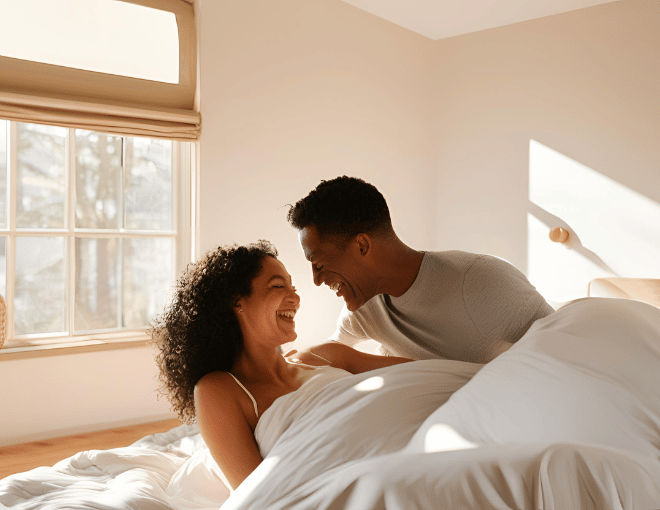 A fit, smiling couple, waking in their sunlit bedroom, stretch comfortably under a soft duvet, with morning light casting gentle shadows across the room.